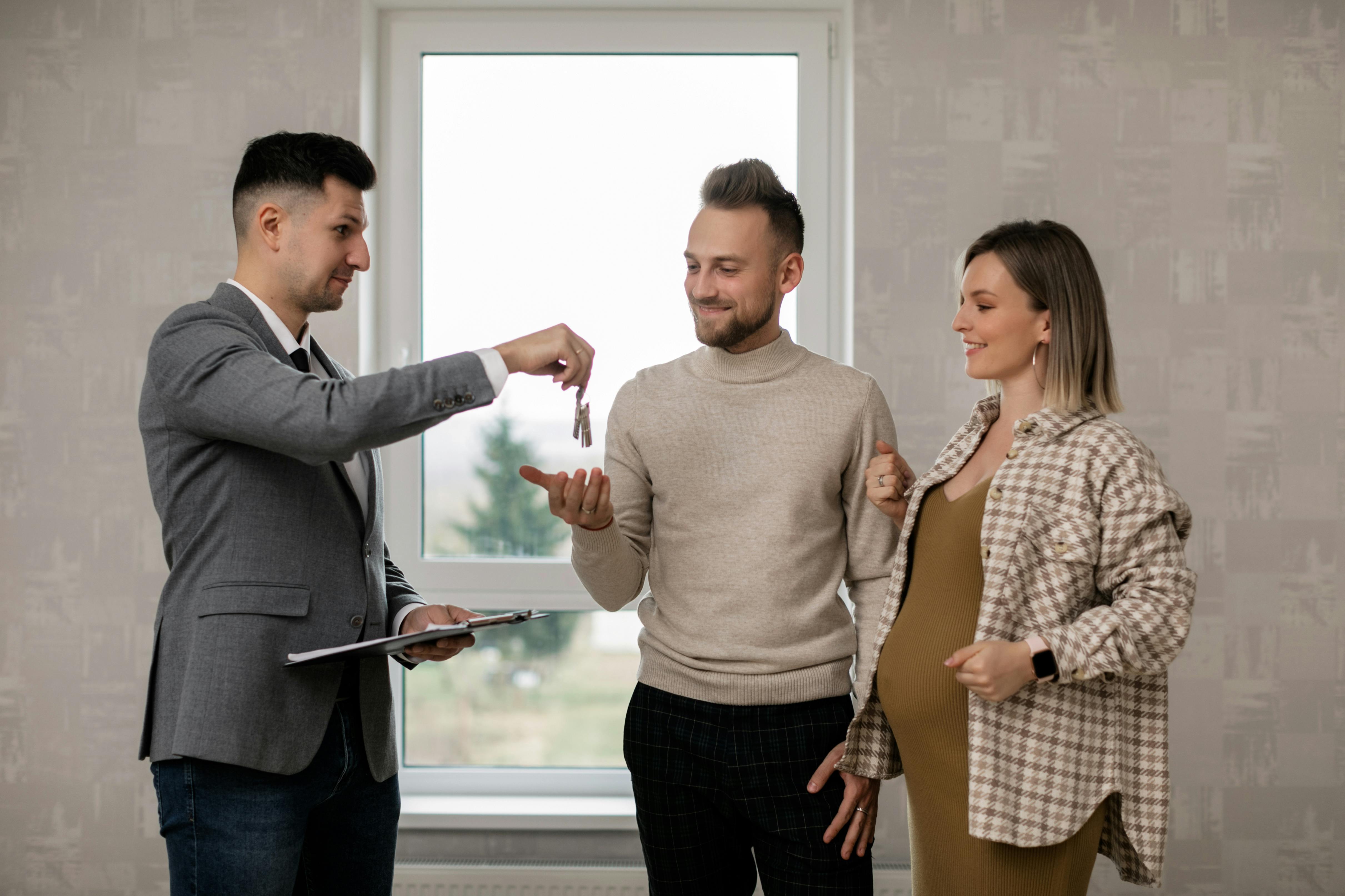 Person handing keys to a smiling couple, with the woman visibly pregnant, standing inside their new home.