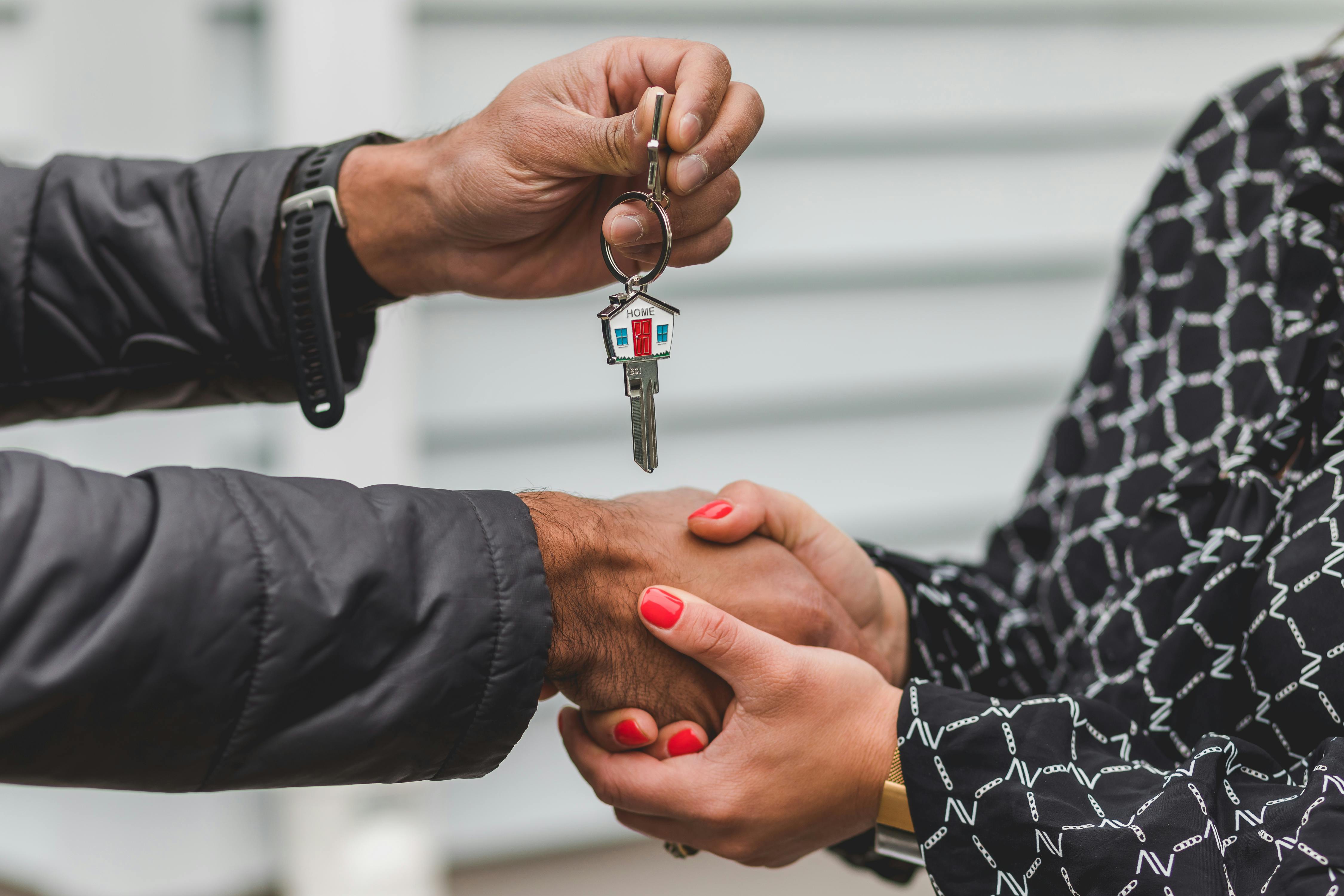 Close-up of a hand giving a keychain with a house-shaped key fob to another hand, symbolizing handing over house keys.