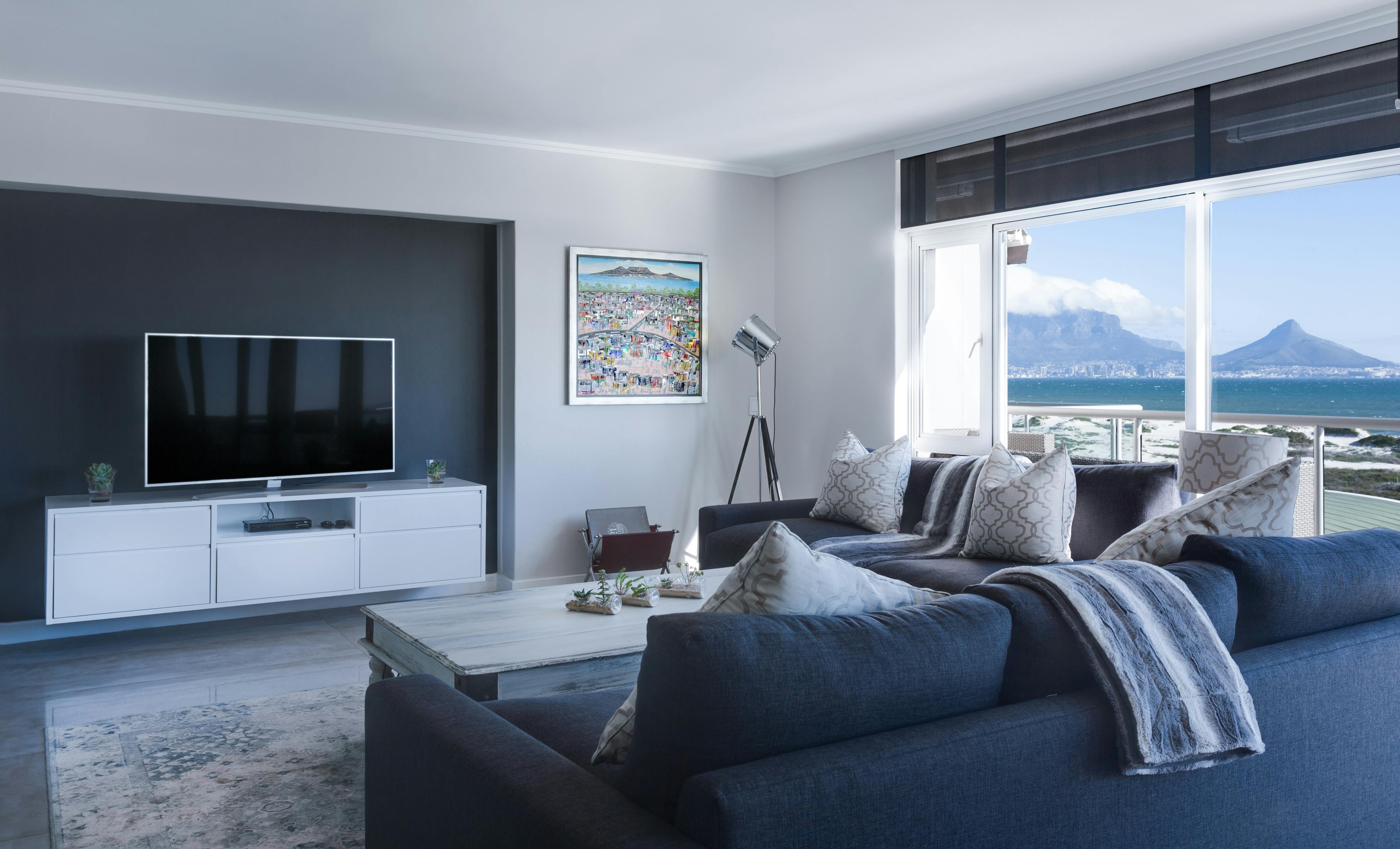 Modern living room with a large window showcasing a scenic mountain and ocean view, featuring a dark blue sofa, a white entertainment unit with a flat-screen TV, and elegant decor.