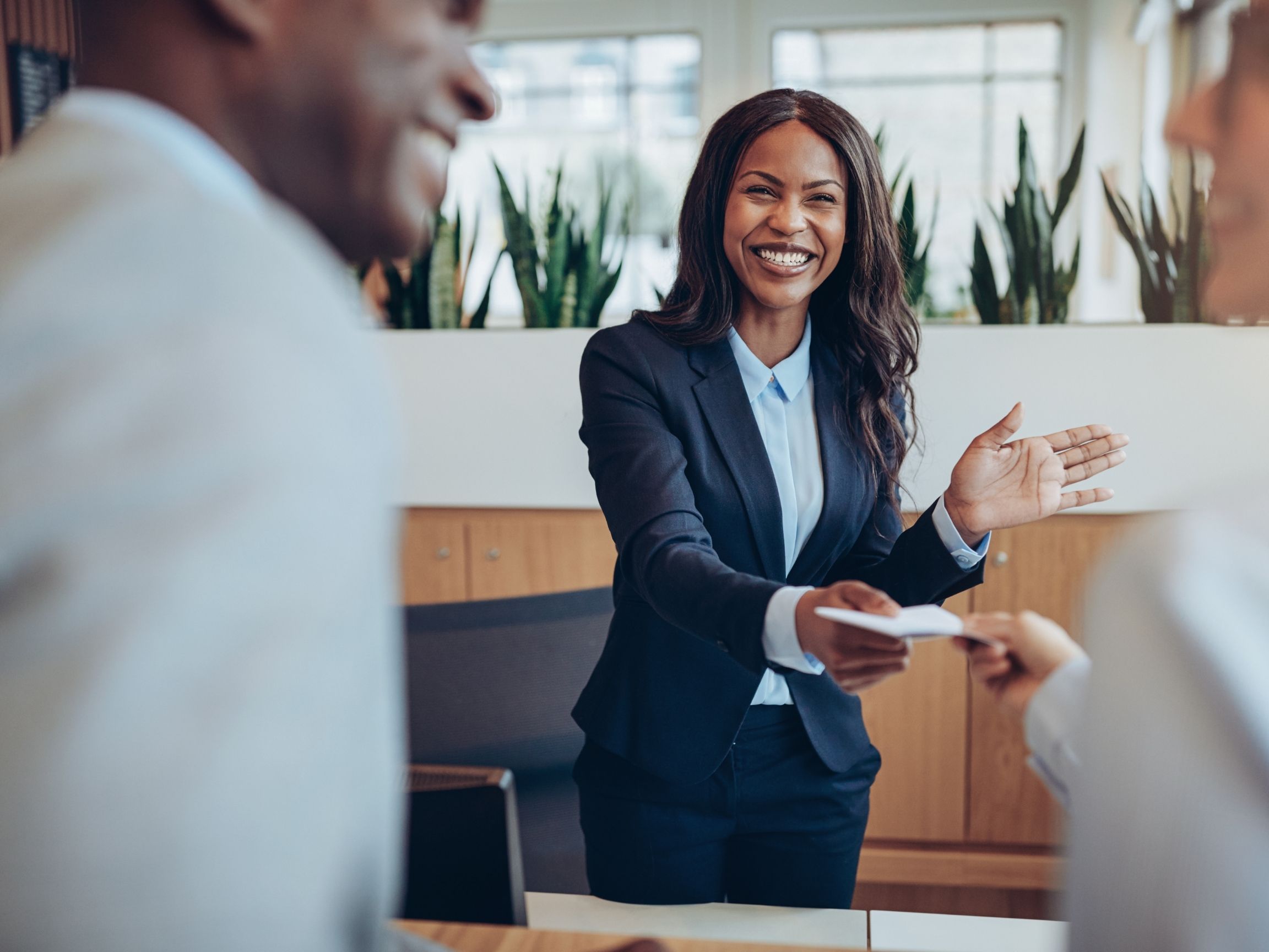 Smiling woman in a suit handing a card to a client, with another client in the foreground.