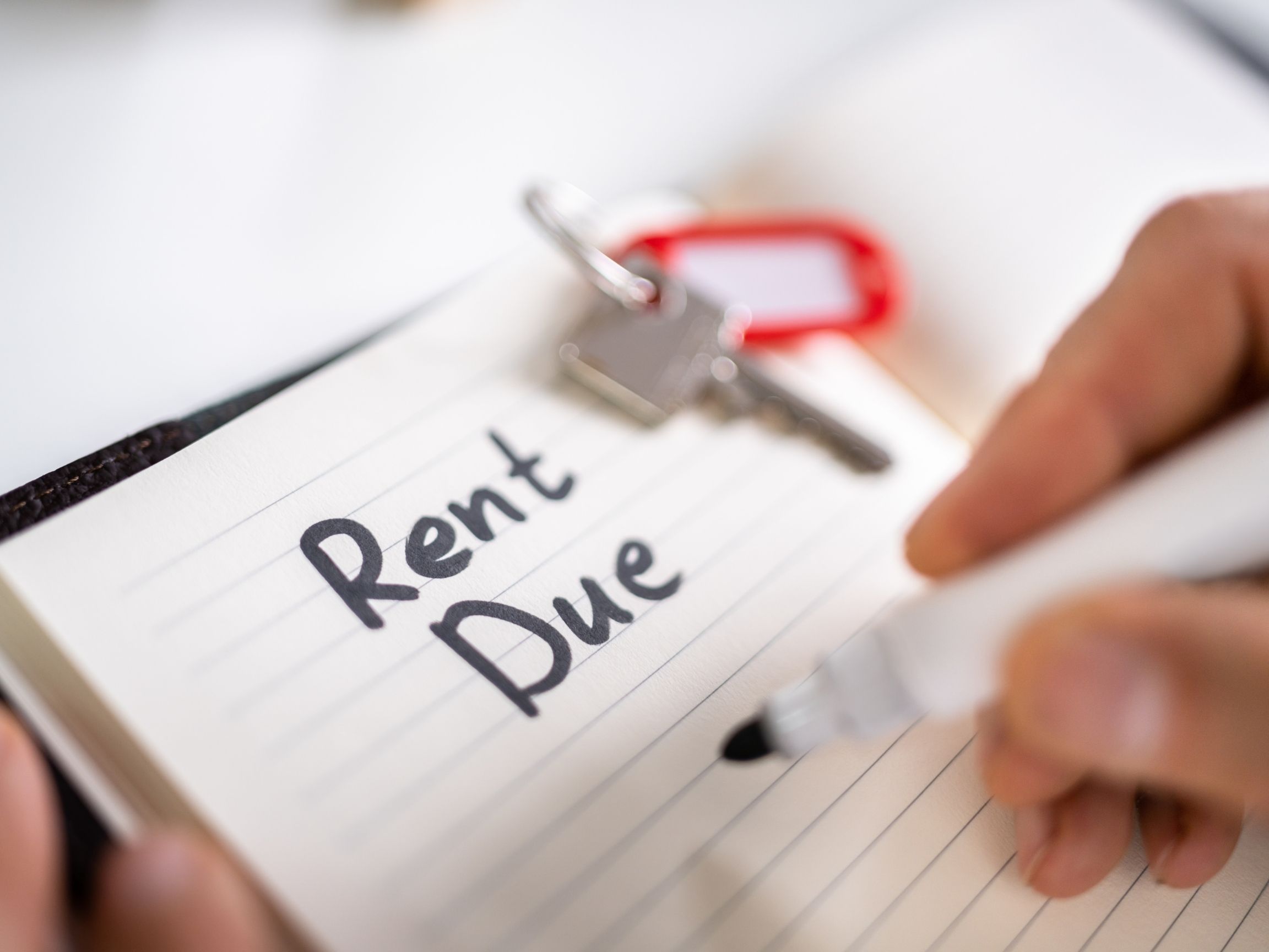 Close-up of a hand writing 'Rent Due' in a notebook with a keychain and keys in the background.