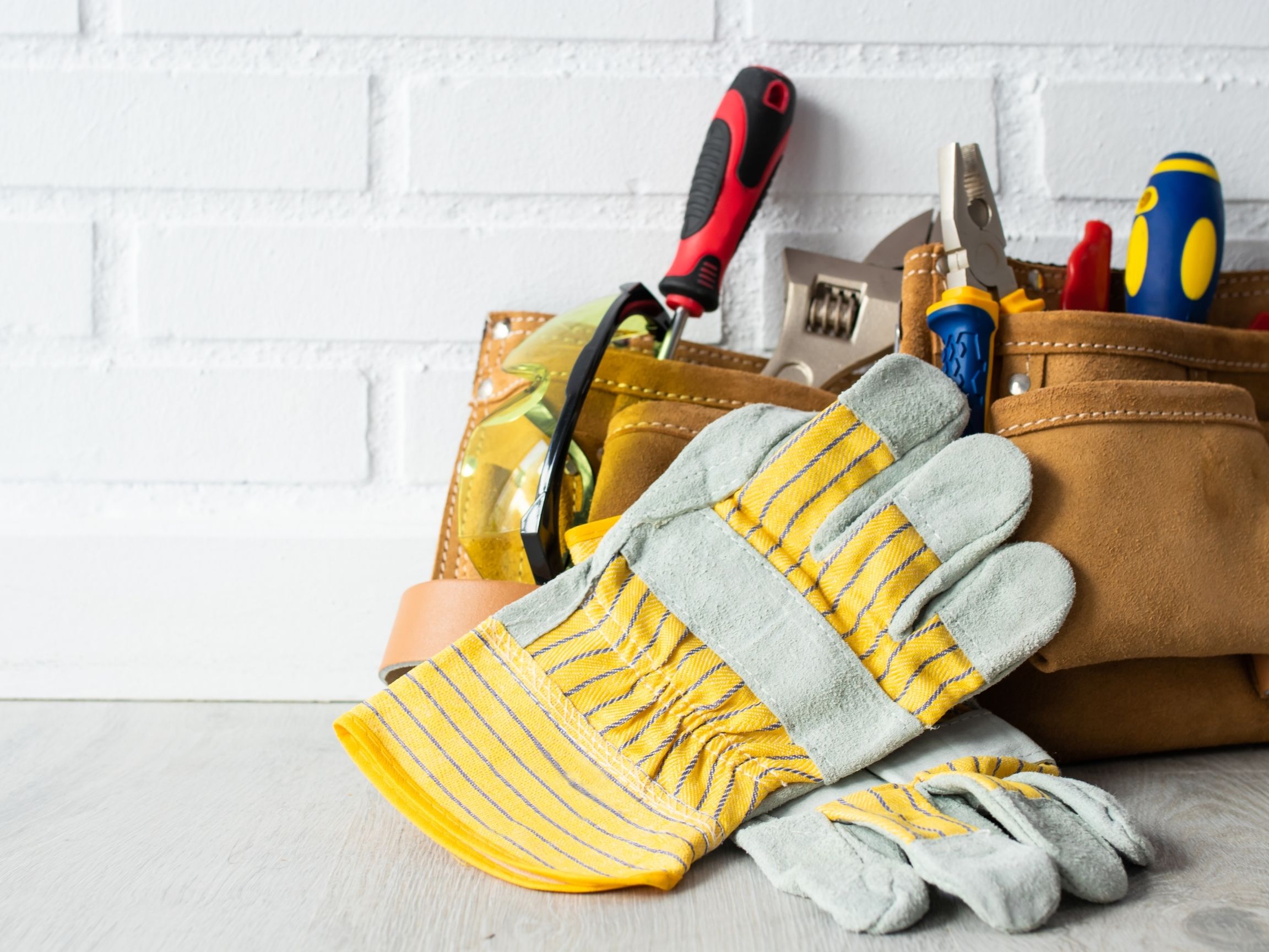 Tool belt filled with various tools and a pair of yellow-striped work gloves placed in front, set against a white brick wall.