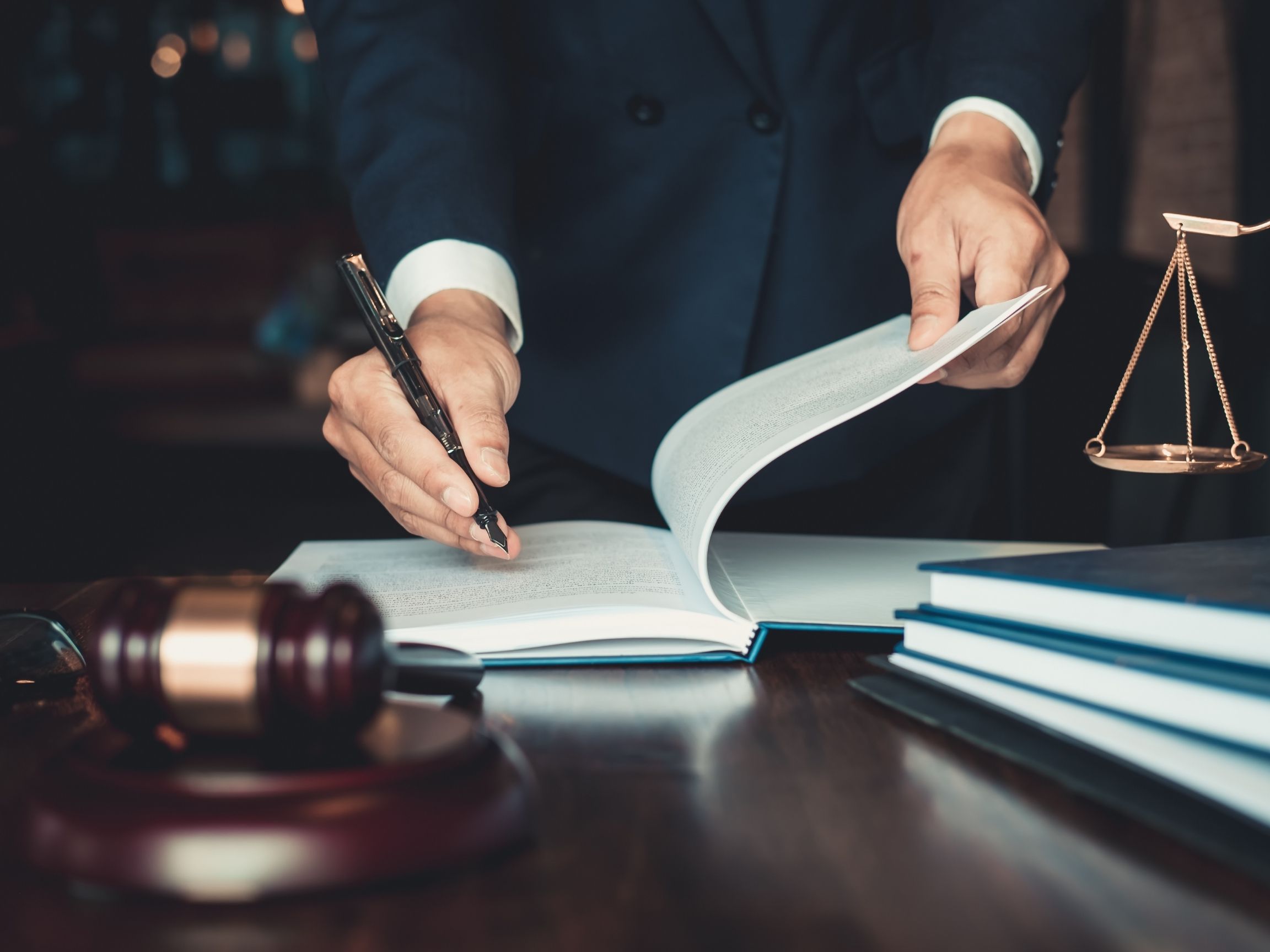 A person in a suit signing legal documents at a desk, with a gavel and scales of justice nearby."