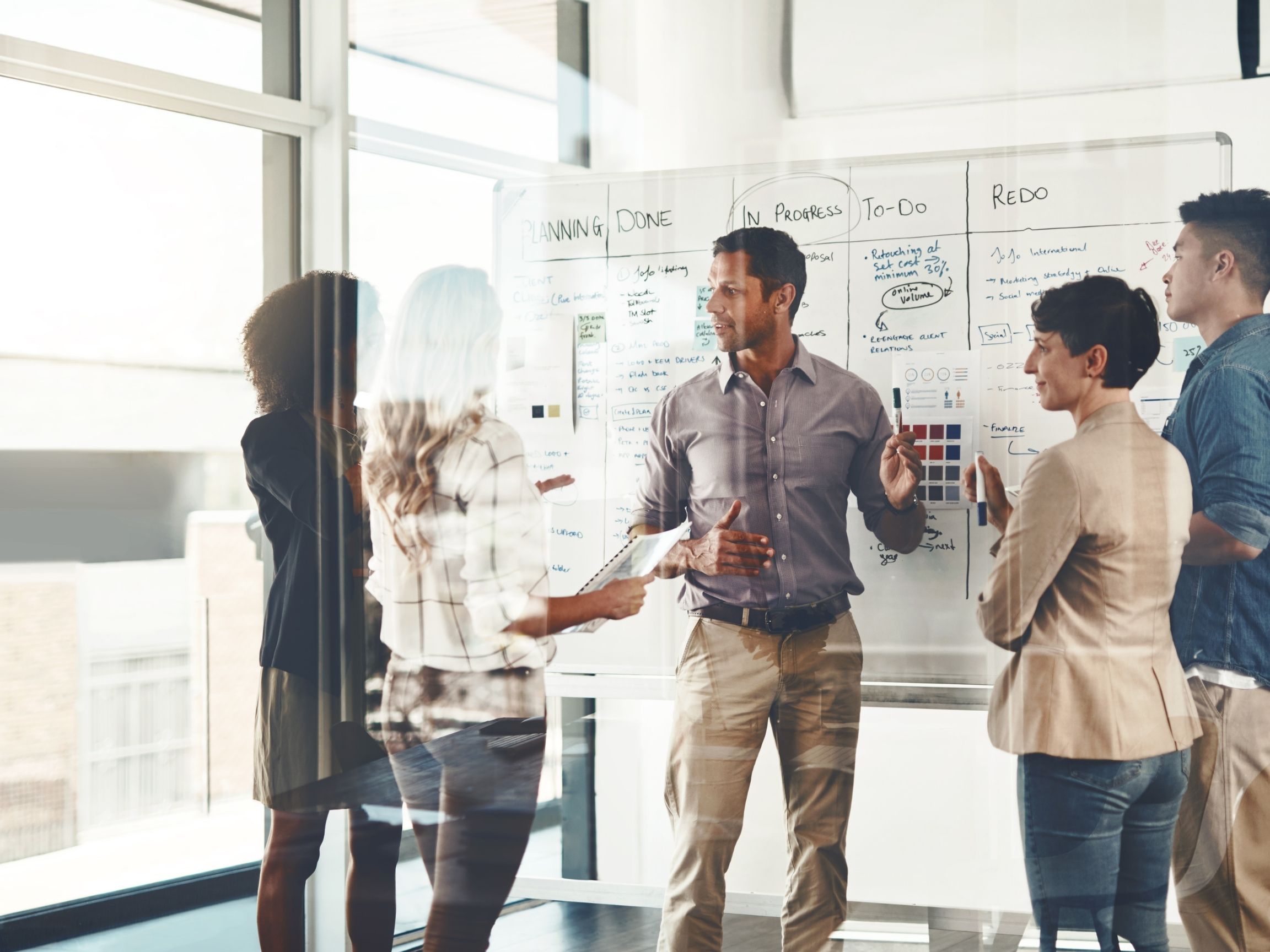 Group of professionals having a meeting in a modern office, standing around a whiteboard with planning and progress notes, seen through a glass wall.