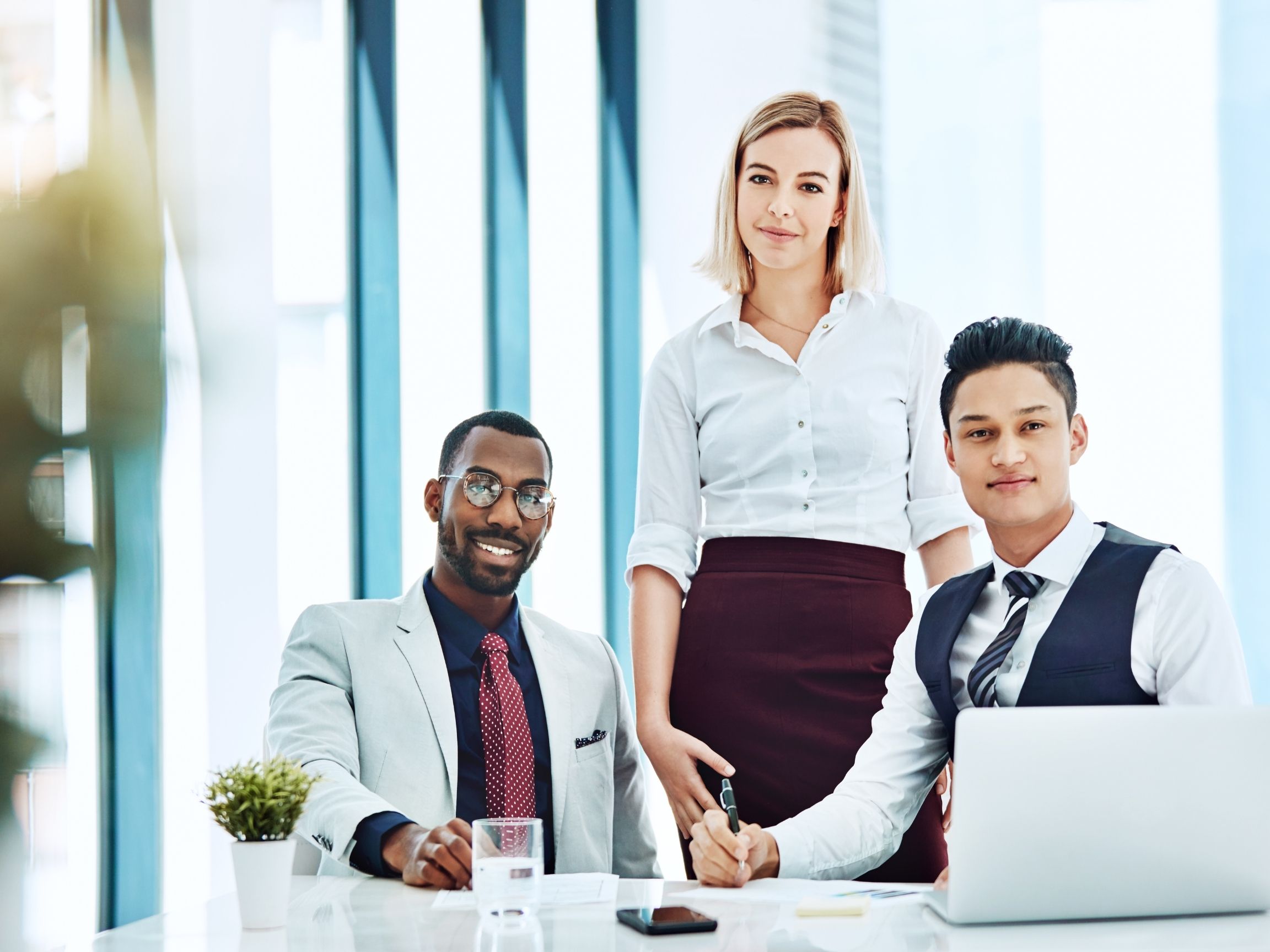 Three business professionals in a modern office setting, with one woman standing and two men sitting at a desk with a laptop and documents.