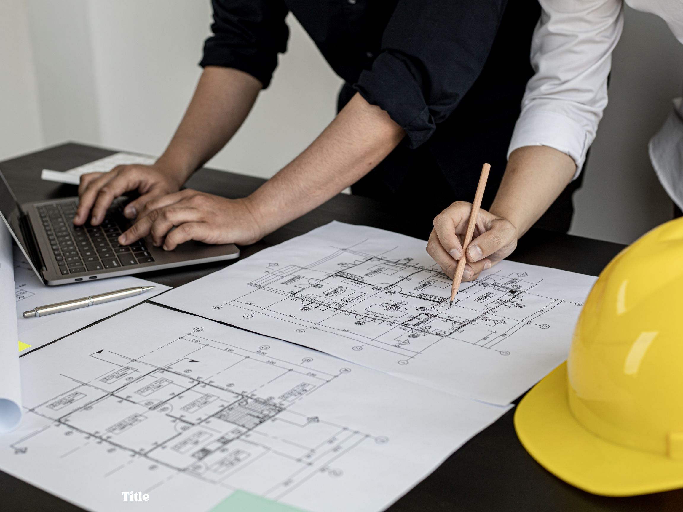 Two people reviewing architectural blueprints on a desk, one working on a laptop and the other holding a pencil, with a yellow hard hat nearby.