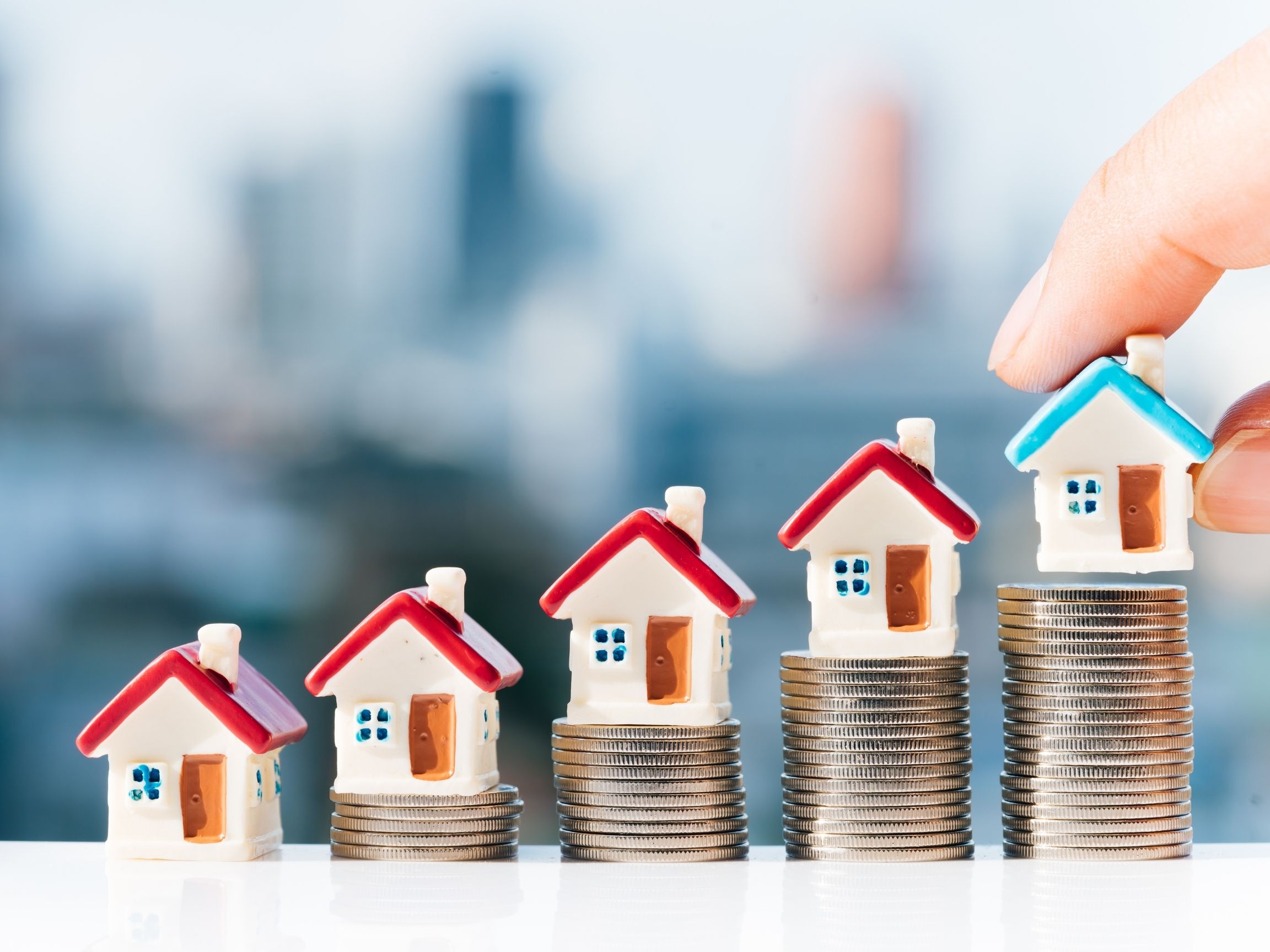 Small model houses placed on stacks of coins, with a hand placing a house on the highest stack, representing the purchasing power of buying multiple properties.